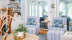 A room with striped chairs, a round table with a plant, a bookshelf, and a view through a glass door. - Goin' Coastal: East Beach 5BR Retreat | Private Pool | Steps to Beach | Sleeps 12 (Saint Simons Island)