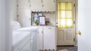 A laundry area with a washing machine, a cabinet with a picture frame and potted plants, and a door with a yellow curtain.