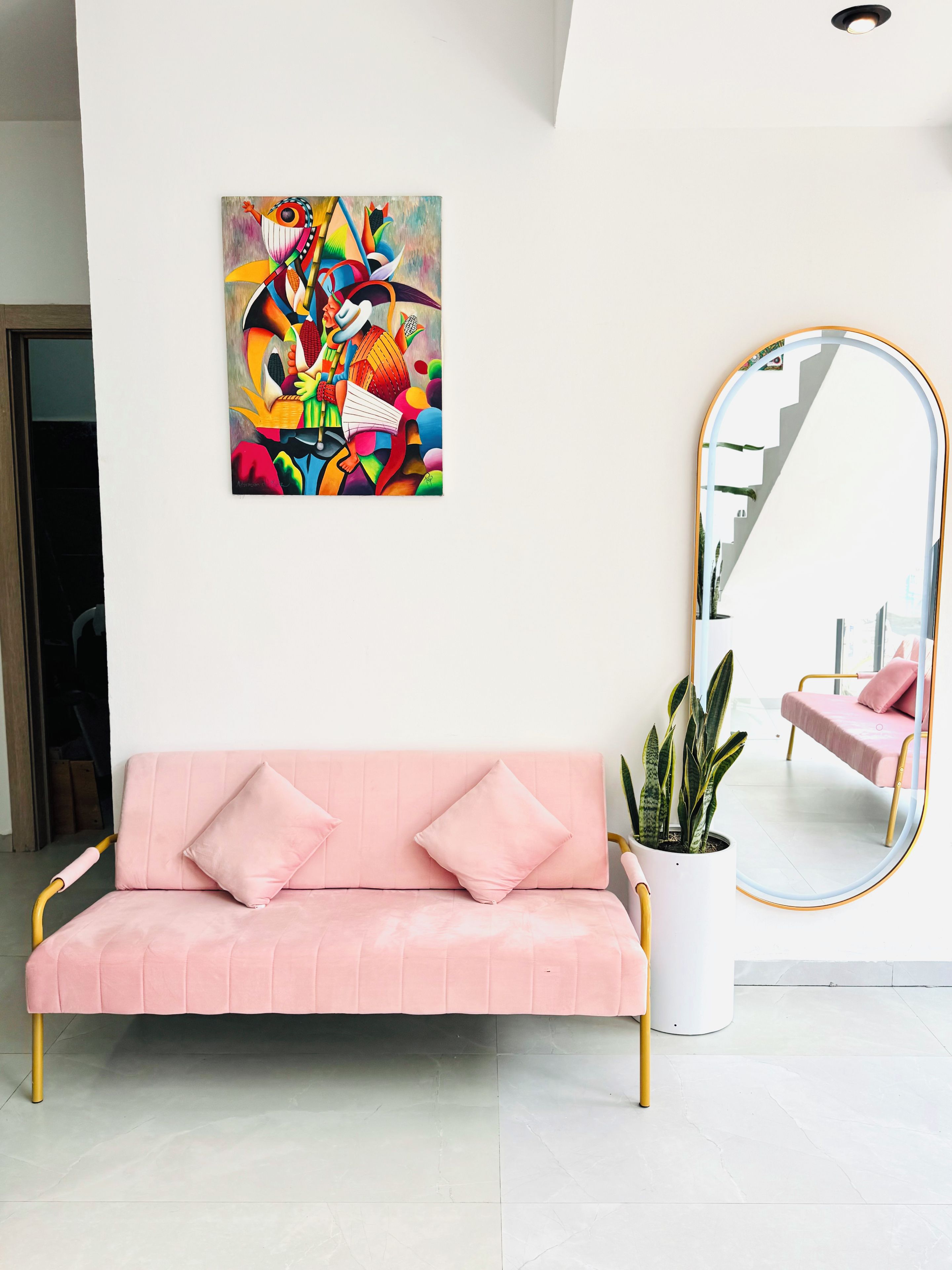Un sofá rosa con cojines, un cuadro abstracto de colores, un espejo grande y una planta en maceta.