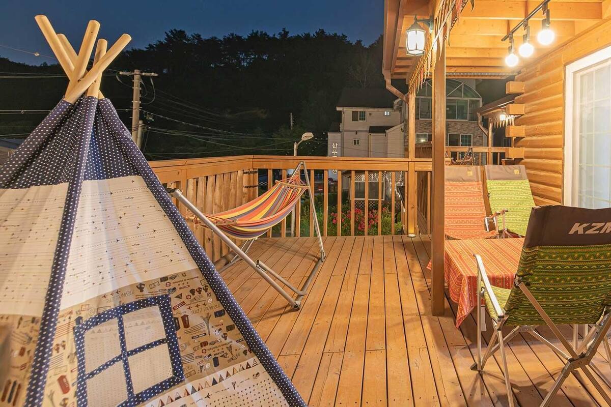 A wooden deck with a hammock, a table with chairs, and a view of a cabin.