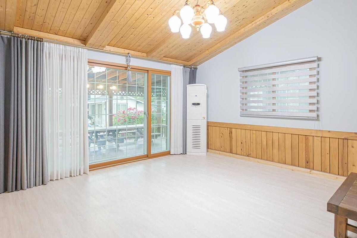 A spacious room with a wooden ceiling, a large sliding glass door leading to an outdoor area, a white air conditioning unit, and a wooden bench.