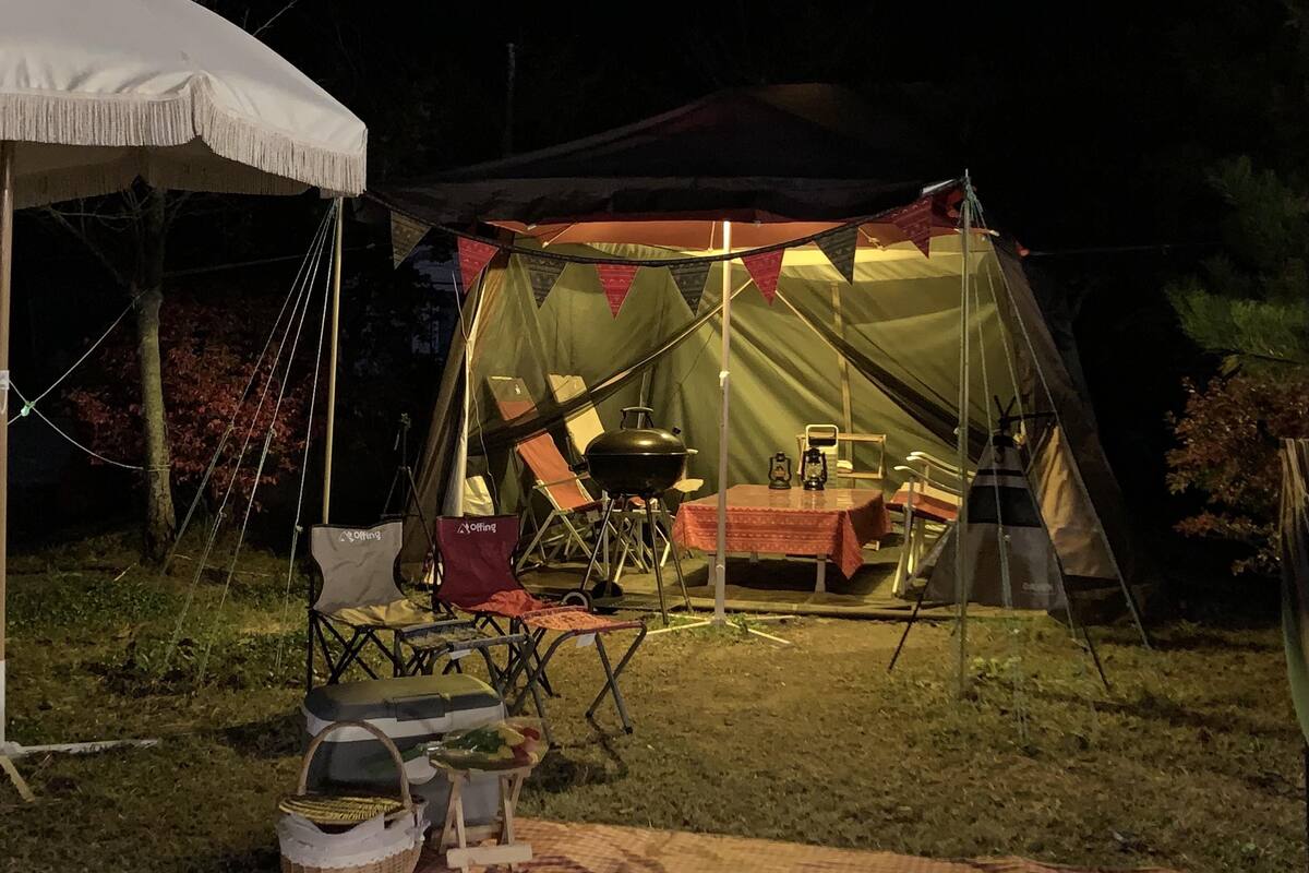 A tent set up in a grassy area at night with a table and chairs inside, a grill outside, and camping chairs and a basket nearby.