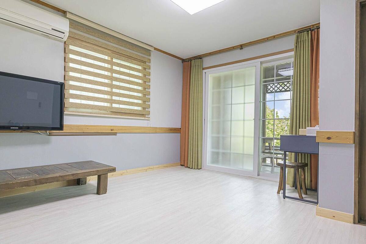 A room with a wooden bench, a television, a table with a chair, and a sliding glass door leading to a balcony.