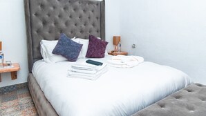 Un lit bien fait, avec une tête de lit capitonnée, des oreillers, une couverture pliée et une table de chevet sur laquelle se trouvent une lampe et une bouteille.