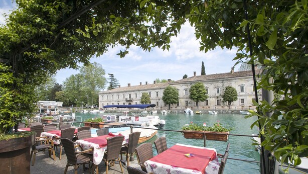 A riverside restaurant with outdoor seating, a view of the water, and historic buildings in the background. - Casa di Paola: Lake-View Balcony Apartment in Peschiera, Peschiera del Garda, Italy (Peschiera del Garda)