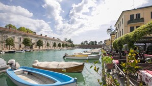 A waterfront area with boats docked along the canal, a row of buildings with balconies, and outdoor seating for cafes. - Casa di Paola: Lake-View Balcony Apartment in Peschiera, Peschiera del Garda, Italy (Peschiera del Garda)