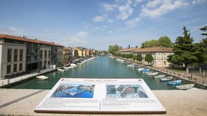 A canal with boats, historic buildings, and a signboard. - Casa di Paola: Lake-View Balcony Apartment in Peschiera, Peschiera del Garda, Italy (Peschiera del Garda)