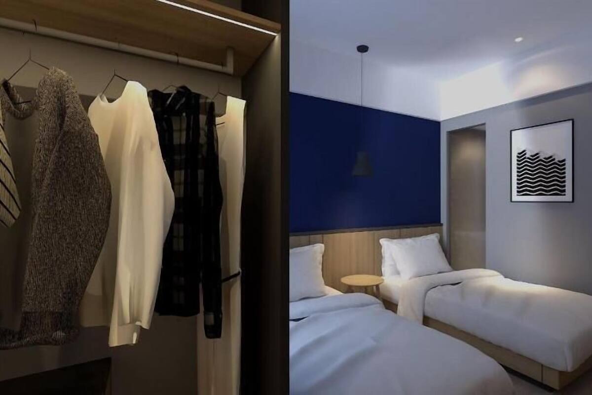 A modern hotel room with a wardrobe, two beds, a small table, and a framed artwork on the wall.