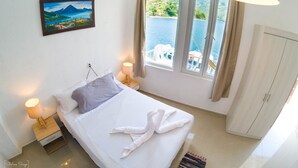 A hotel room with a bed, a painting, a window with a view of the sea, and a wardrobe. - San Pedro Suite (San pedro la Laguna solola)