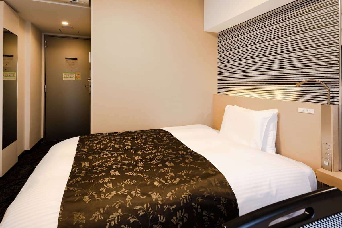 Een hotelkamer met een bed, een hoofdeinde met een bedieningspaneel, een raam met jaloezieën en een airconditioning.