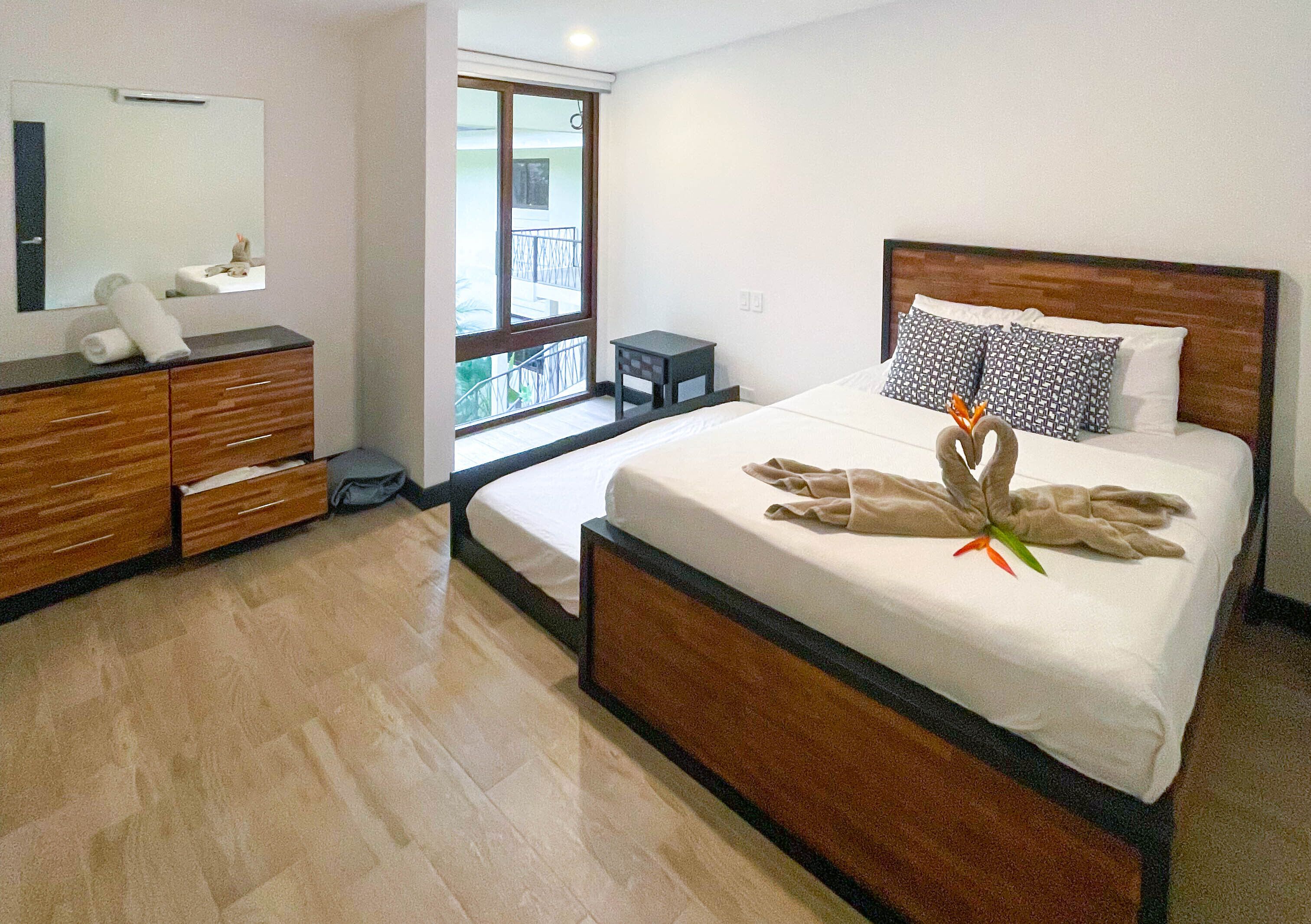 A hotel room with a wooden bed, a bedside table, a mirror, a dresser, and a balcony view.
