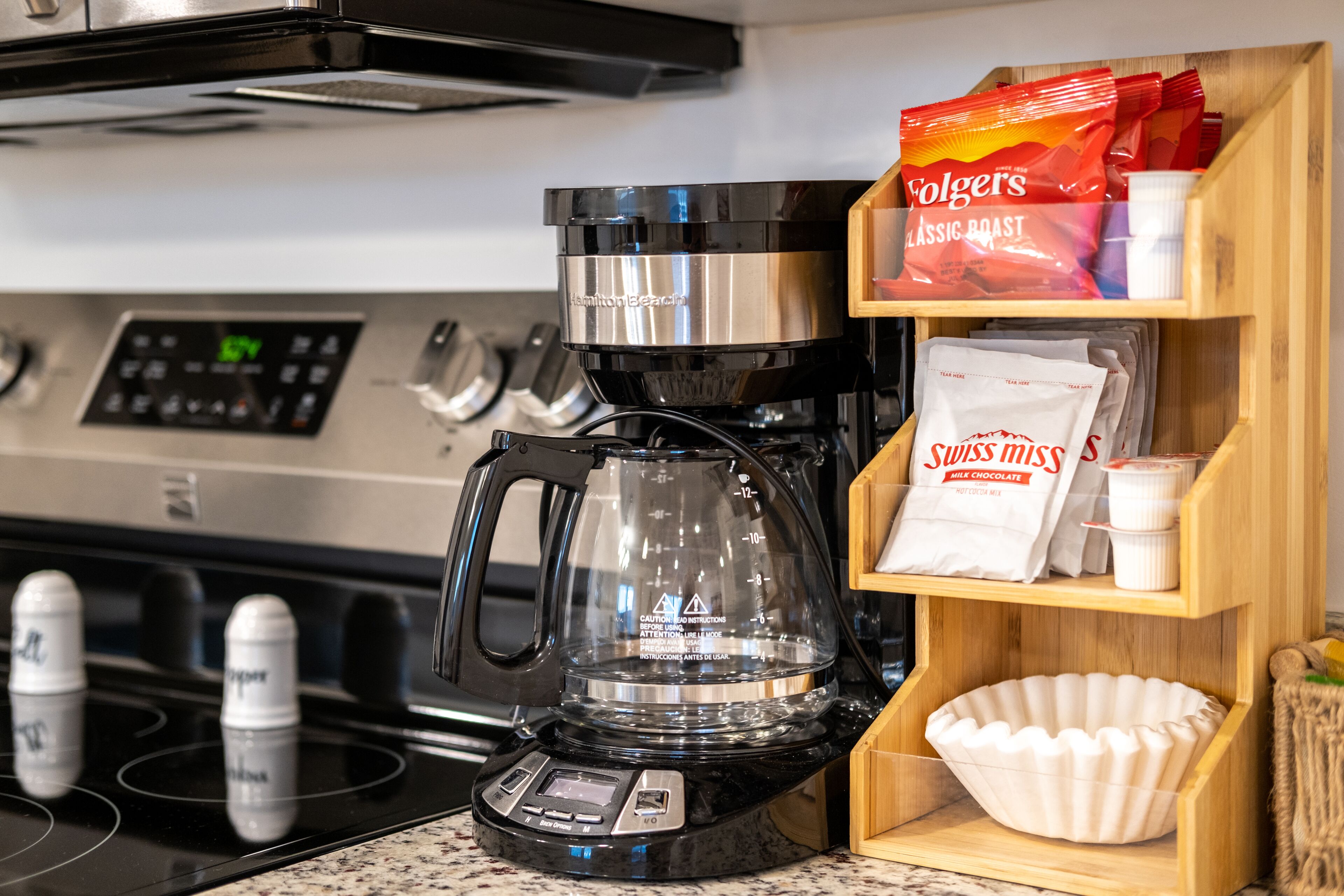 Une cuisine équipée d’une machine à café, d’une cuisinière et d’étagères où sont rangées des dosettes et des paquets de café.