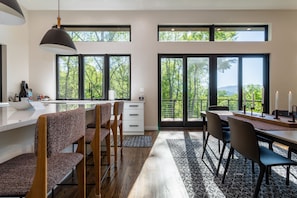 A modern kitchen with a large island, a dining area with a table and chairs, and large windows offering a view of trees and mountains.