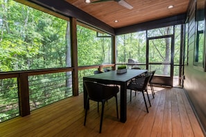 A wooden deck with a table and chairs, large windows offering a forest view, and a ceiling fan.