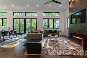 A spacious living room with a fireplace, large windows, and a modern ceiling fan.