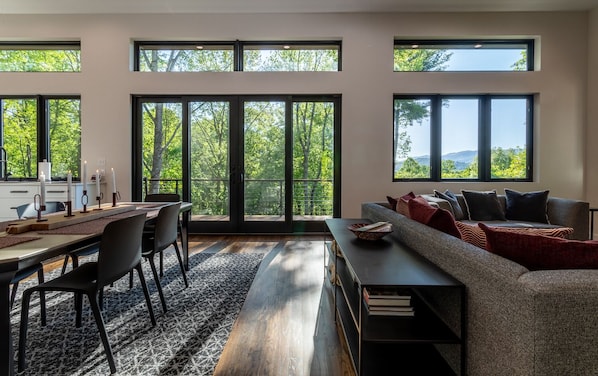 A modern living room with a dining area, large windows, and a view of trees.