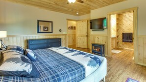 A bedroom with a bed, wooden ceiling, a ceiling fan, a TV, and a fireplace.