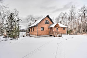 Une cabane en bois dotée d’un porche et d’une cheminée, entourée de neige et d’arbres.