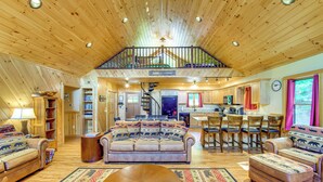 Ein geräumiger Wohnbereich mit hohen Decken, Holzböden, einer zentralen Sitzecke mit Sofa und Hocker, einem Esstisch mit Stühlen, einer Küche im Hintergrund und einer Treppe, die zu einer oberen Ebene führt.