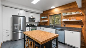 Une cuisine moderne avec des meubles blancs, des appareils en acier inoxydable, un îlot central en bois avec des tabourets et une fenêtre donnant sur les arbres.