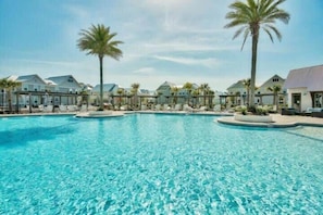 En stor, klar swimmingpool omgivet af liggestole og palmer, med resort-lignende bygninger i baggrunden.