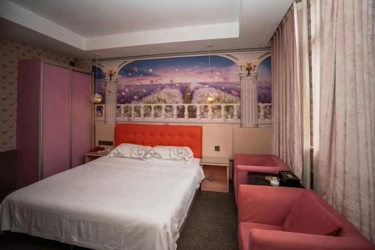 A hotel room with a bed, a red upholstered chair, a nightstand, and a mural on the wall.
