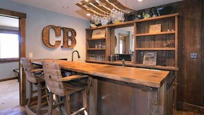 Zona de bar con mostrador de madera, taburetes y una estantería con vasos y botellas.