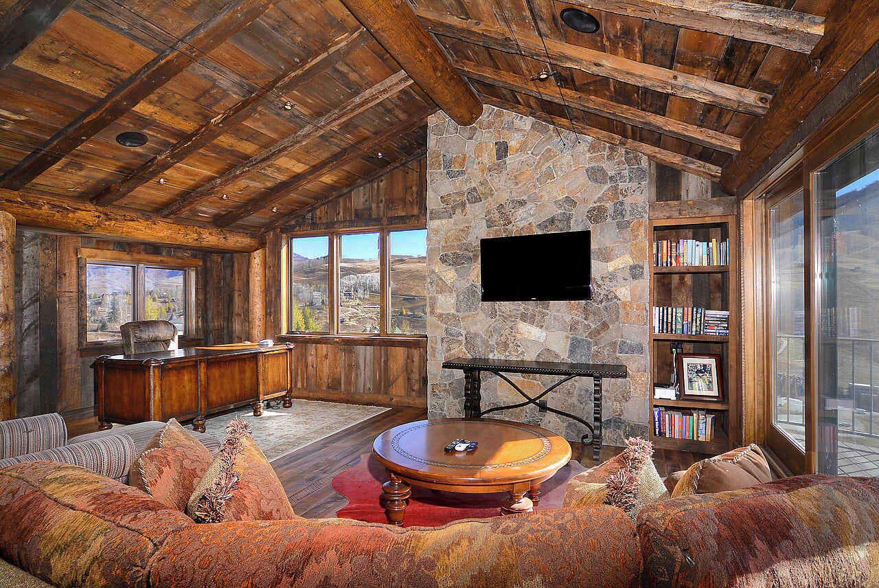 A rustic interior with a stone wall, wooden ceiling, a flat-screen TV, a bookshelf, and a large window.