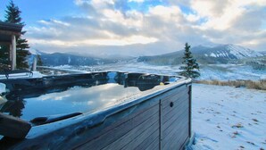 Un jacuzzi con revestimiento de madera, rodeado de nieve y montañas al fondo.