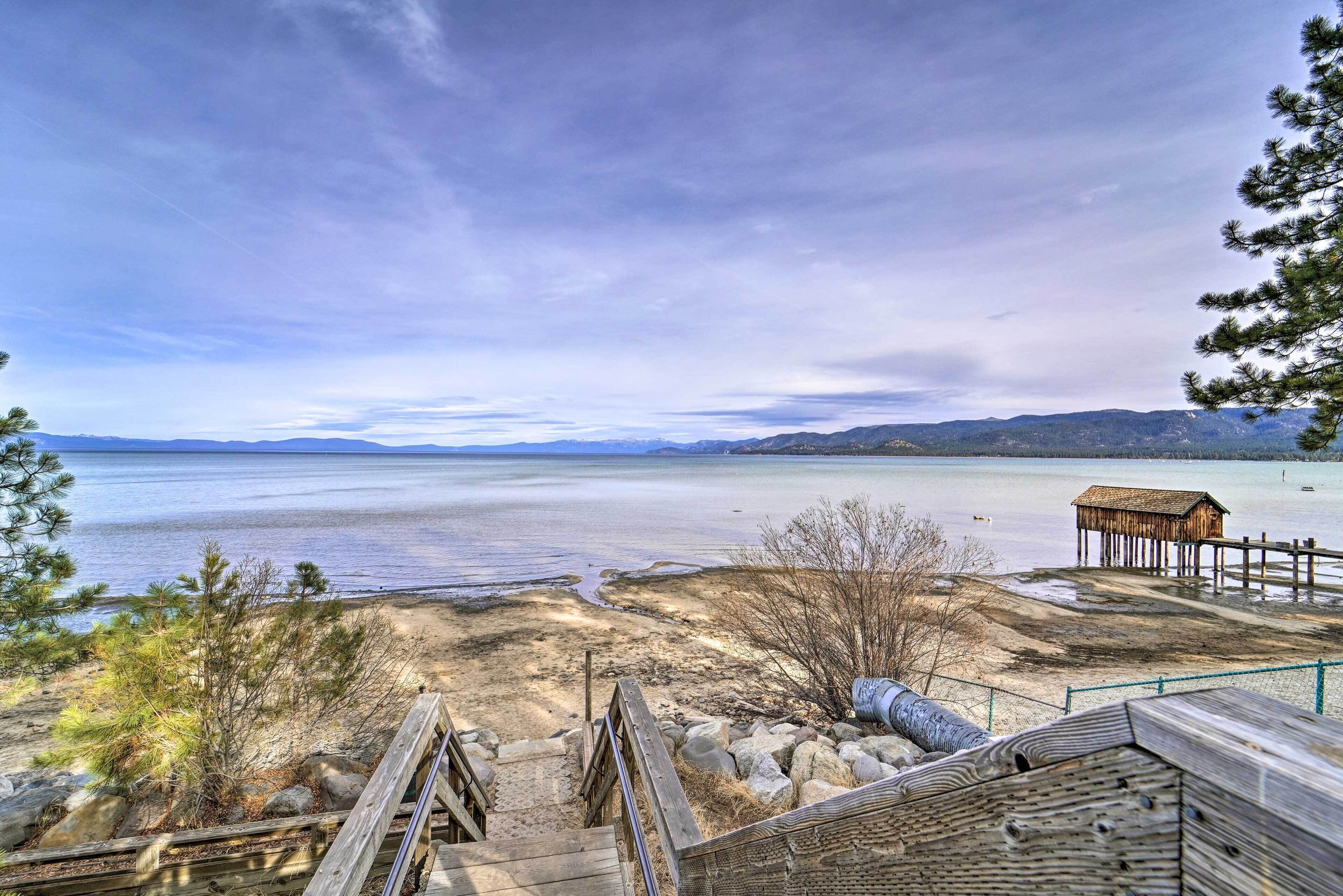 A wooden deck leading to a beach with a thatched-roof structure in the distance.