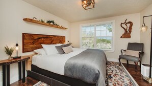 A bedroom with a large bed, a wooden headboard, a window with a view, a chair, and a floor lamp. - Charming Hilltop Getaway - Valley and Water Views (Sequim)