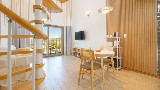 Quarto de hotel moderno com escada de madeira, área de jantar com mesa e cadeiras, televisão de tela plana na parede e uma grande janela com vista para o exterior.