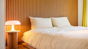 Cama bem arrumada com roupa de cama e almofadas brancas, luminária de cabeceira e cabeceira de ripas de madeira.