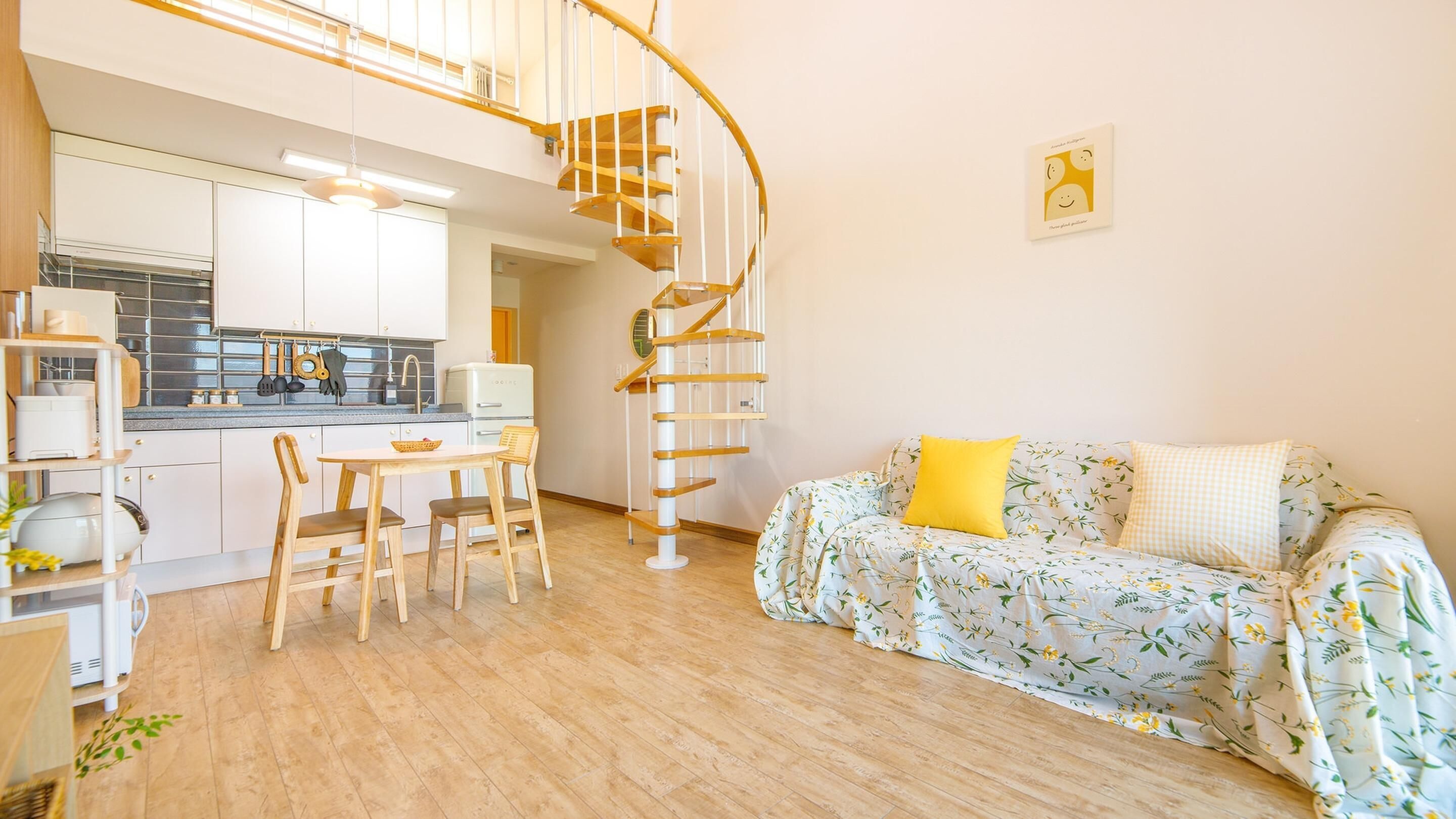 Et moderne køkken med hvide skabe, et træspisebord og stole. En spiraltrappe fører op til et højere niveau. I opholdsstuen er der en sofa med blomstermønster og gule puder.