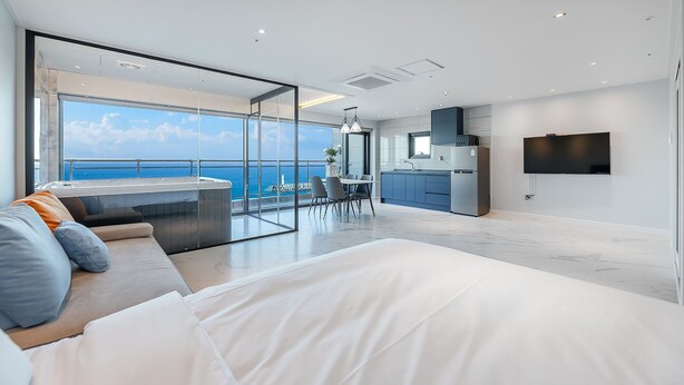 A modern bedroom with a large bed, a balcony with a view of the sea, a kitchen area, and a TV mounted on the wall.
