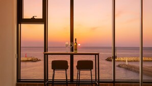 A room with a large window overlooking a sunset over the sea, a table with two chairs, and a bottle of wine.