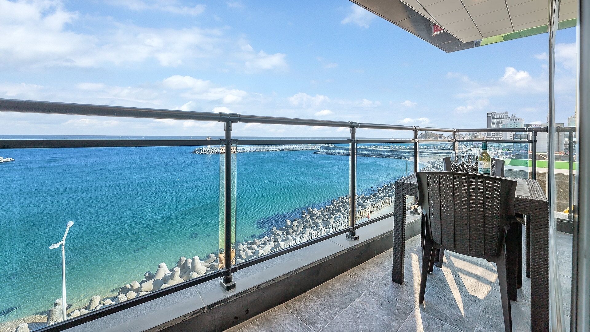 A balcony with a view of the sea, a chair, and a table with a bottle.