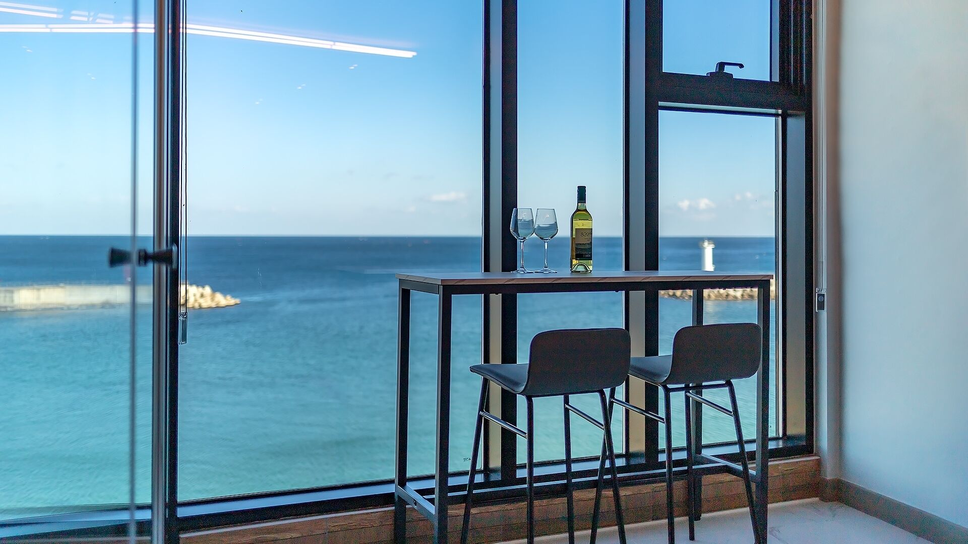 A room with a view of the sea, a table with two chairs, and a bottle of wine.