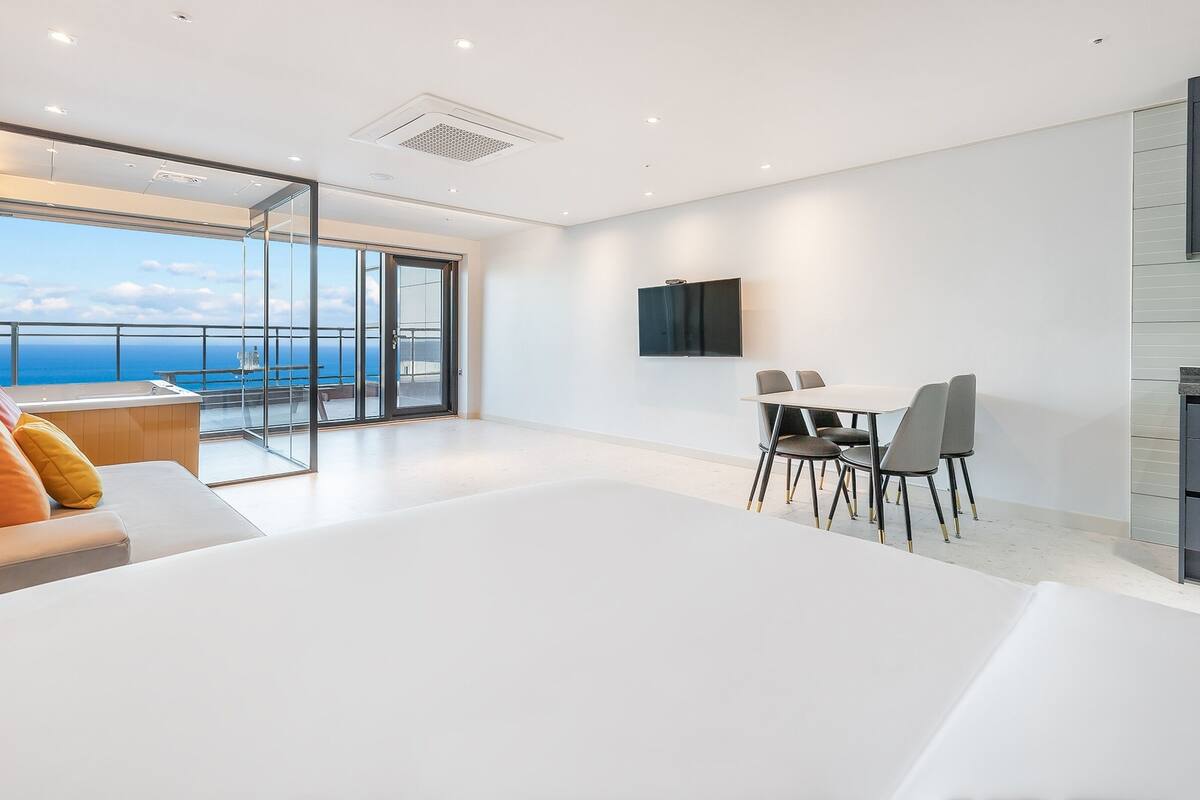 Una habitación de hotel moderna con una cama grande, un comedor con sillas, un televisor montado en la pared y vistas al mar a través de amplias ventanas.