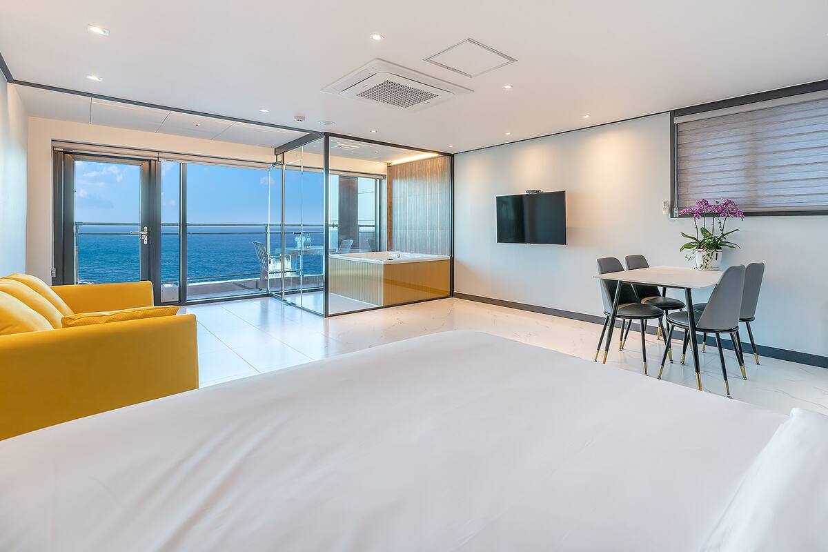 Una habitación de hotel moderna con una cama grande, un balcón con vistas al mar, un comedor con mesa y sillas, y un televisor montado en la pared.