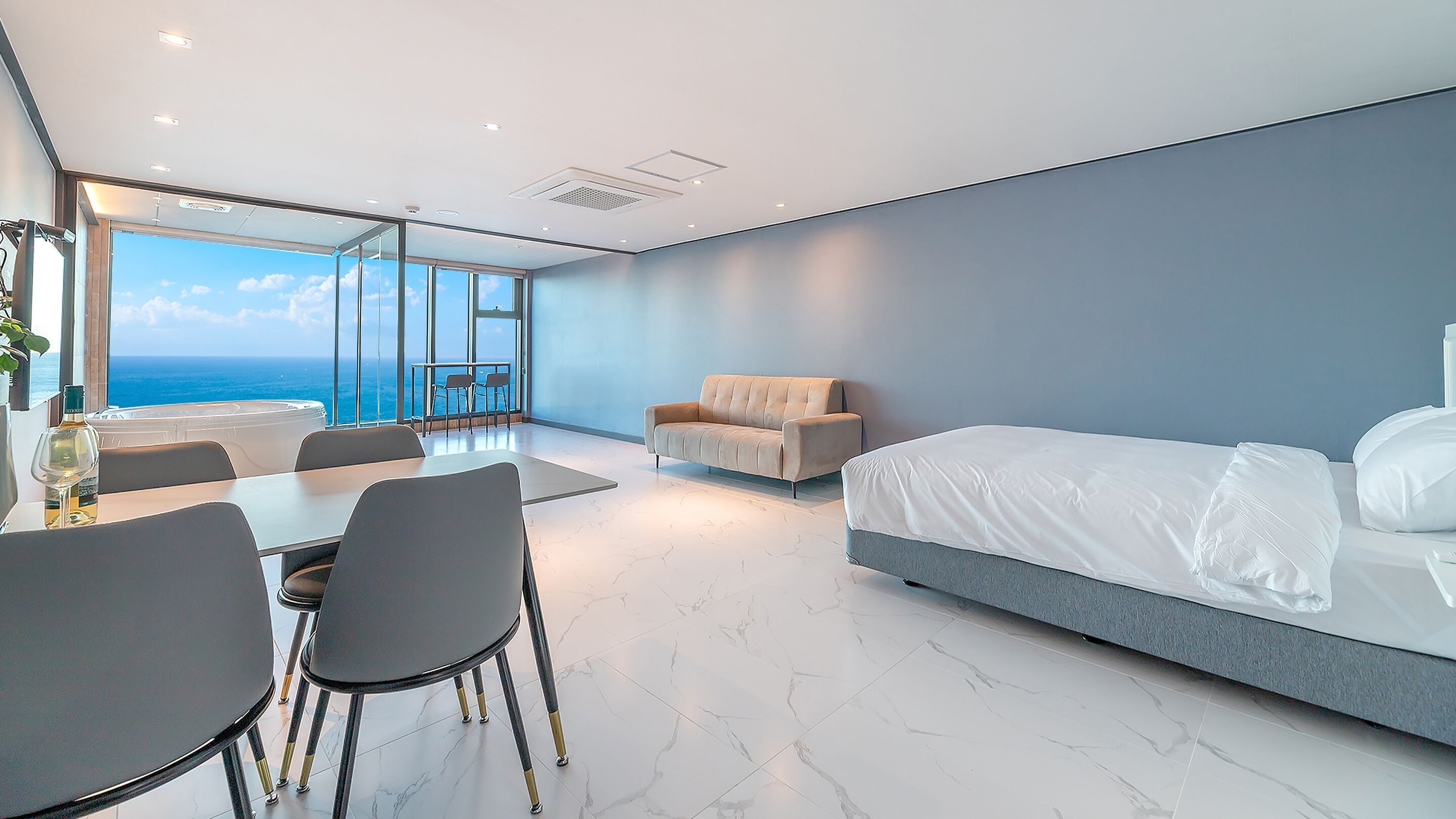 A modern hotel room with a large bed, a sofa, a dining area with a table and chairs, and a view of the ocean through large windows.