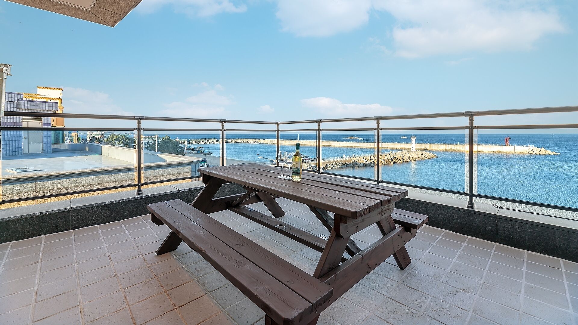 A balcony with a wooden picnic table and a bottle of wine, overlooking a coastal area with a lighthouse and rocky breakwater.