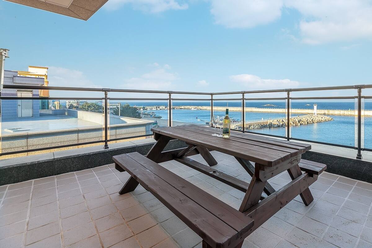 Un balcón con una mesa de picnic de madera y una botella de vino, con vistas a una zona costera que incluye un faro y un rompeolas rocoso.