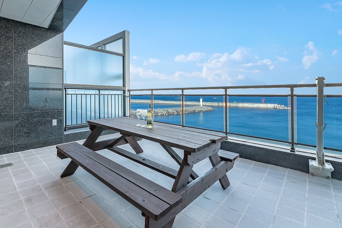 Un balcon avec une table et des chaises en bois, une bouteille de vin et une vue sur la mer.