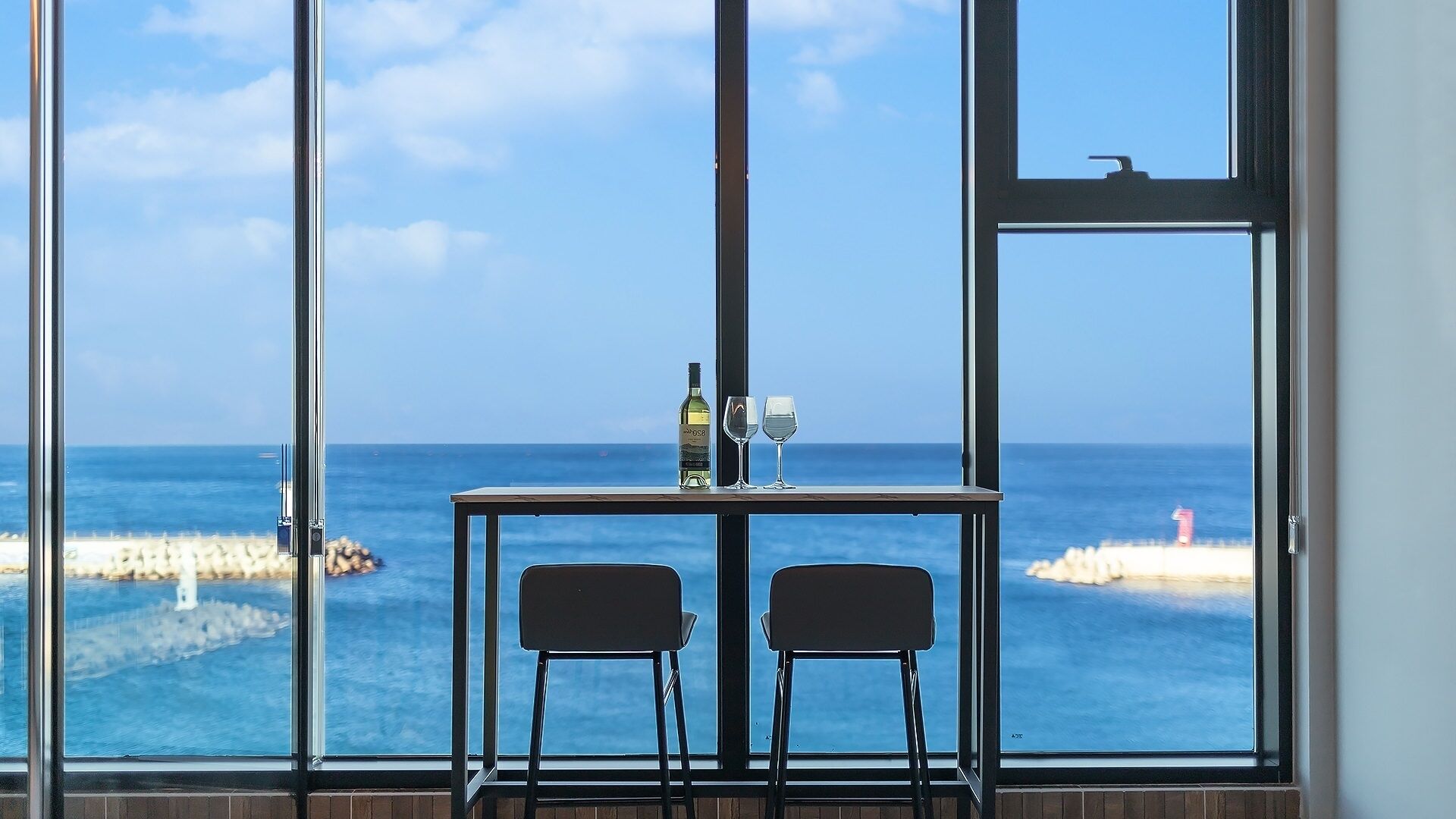 A room with a large window overlooking the sea, a table with two chairs, and a bottle and glasses on the table.