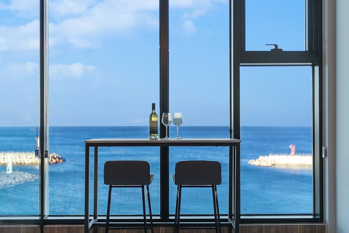 Una habitación con un amplio ventanal con vistas al mar, una mesa con dos sillas y una botella y vasos sobre la mesa.