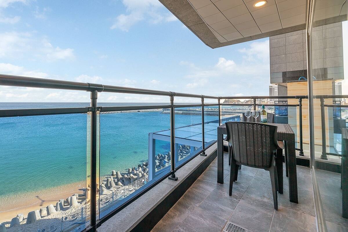 Un balcón con vista al mar, barandilla de cristal y una mesa con sillas.