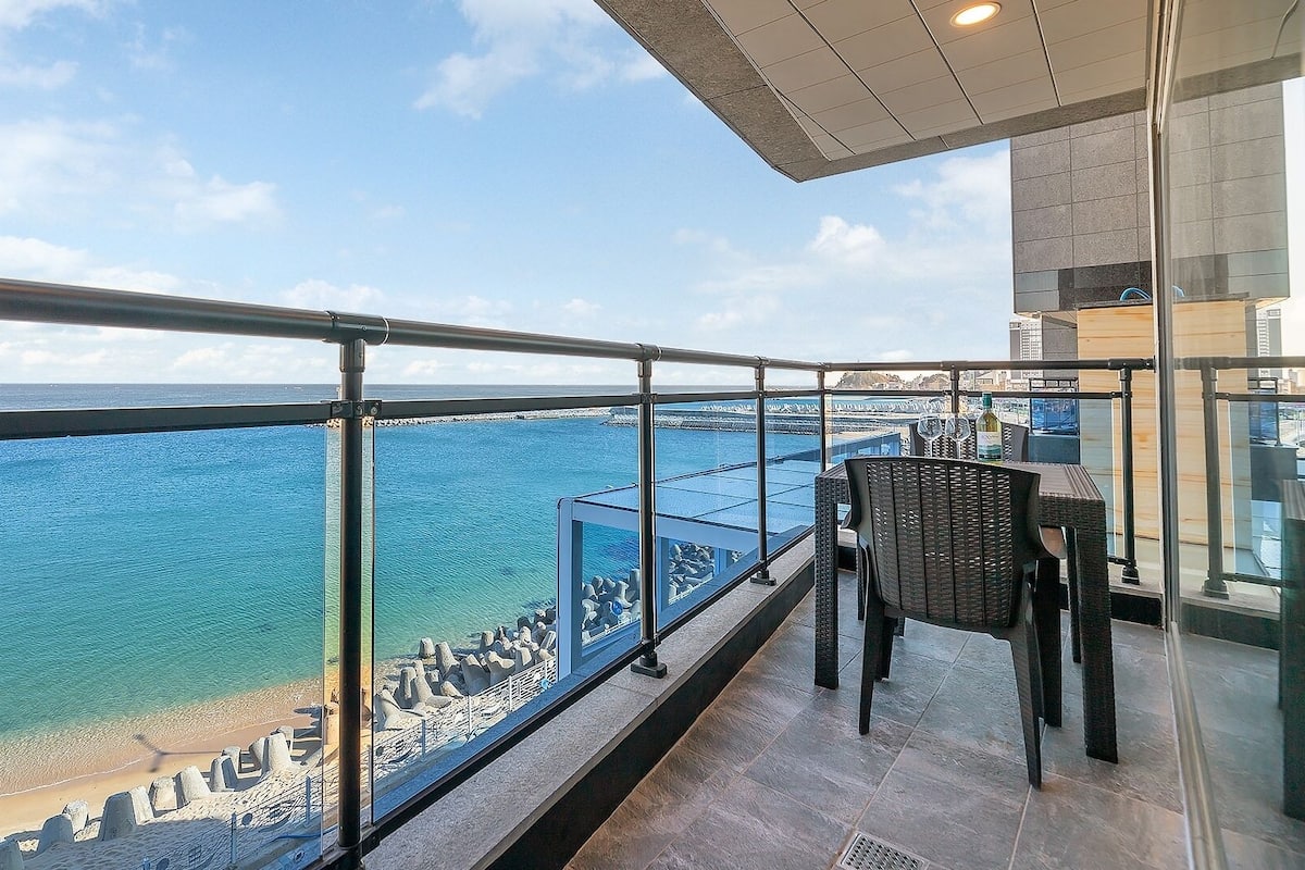 Un balcon avec vue sur la mer, une rambarde en verre et une table avec des chaises.
