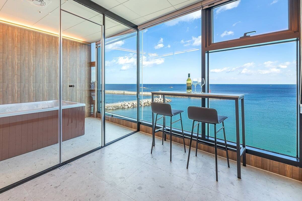 Un baño moderno con separador de vidrio, tocador de madera y una ventana grande con vista al mar.