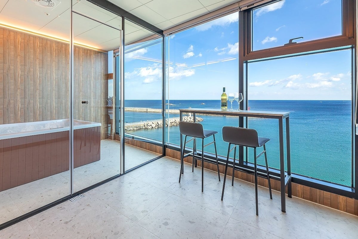 Une salle de bain moderne avec une paroi de verre, un meuble-lavabo en bois et une grande fenêtre donnant sur la mer.